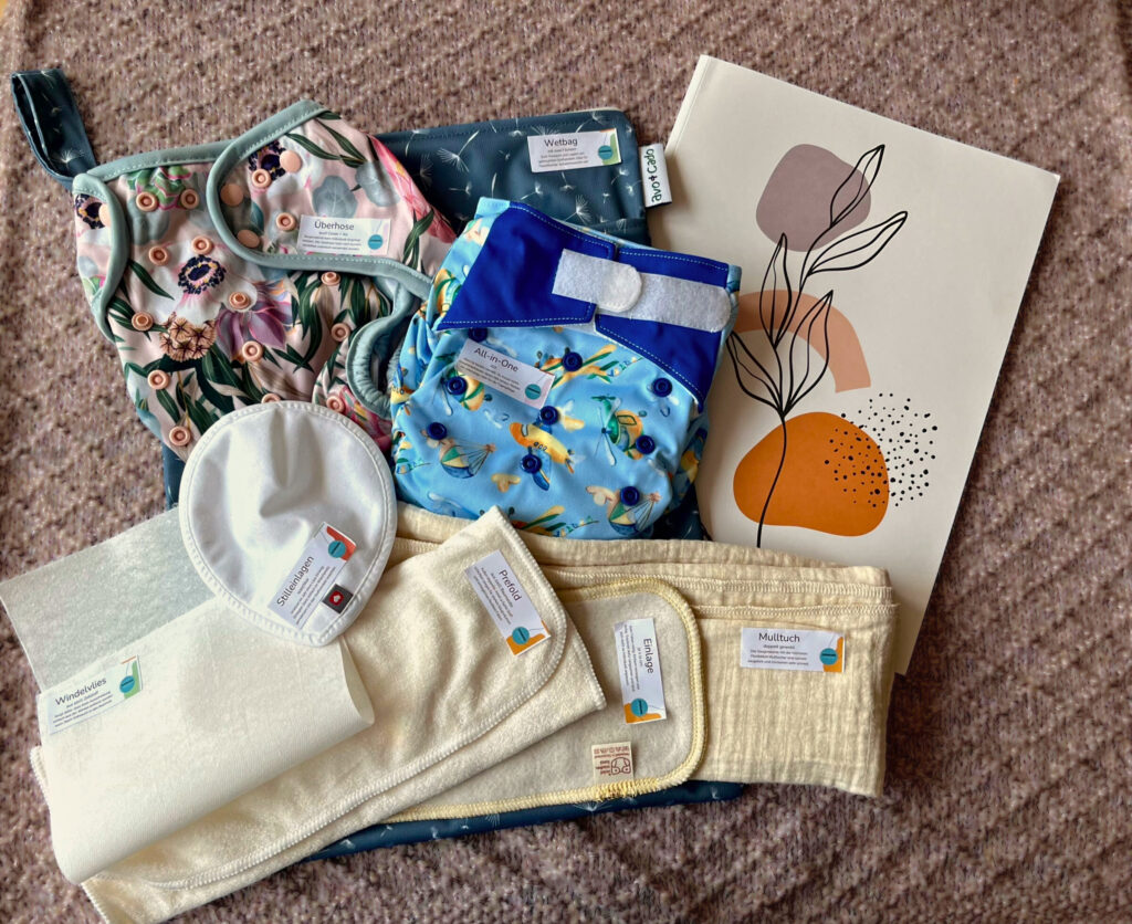 Ein liebevoll zusammengestelltes Stoffwindel-Set liegt auf einer rosafarbenen Decke. Zu sehen sind verschiedene moderne Stoffwindeln in bunten Mustern, ein Wetbag, Einlagen, ein Mulltuch, Stilleinlagen und Windelvlies – alle mit kleinen Etiketten versehen. Rechts daneben liegt ein Poster mit abstrakten Formen und einer Pflanze als Linie gezeichnet.