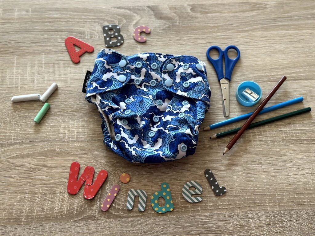 Blau gemusterte Stoffwindel liegt auf einem Holztisch, umgeben von Schulutensilien und bunten Buchstaben