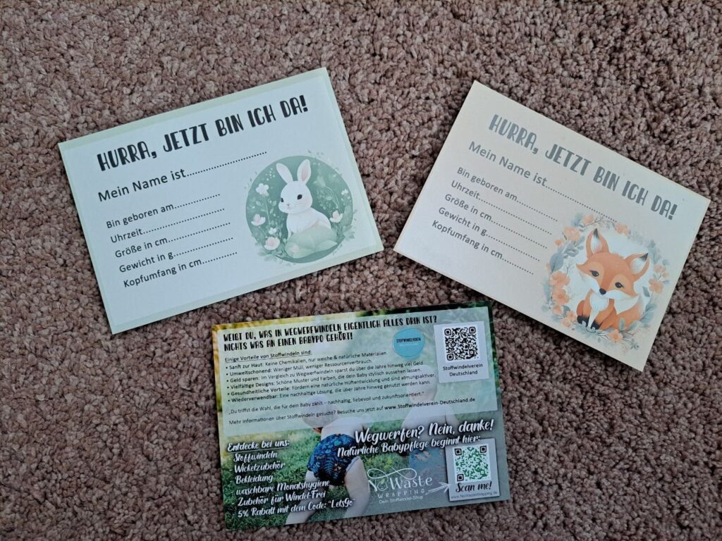 Drei Karten liegen auf einem beigen Teppich. Zwei Karten tragen die Überschrift „Hurra, jetzt bin ich da!“ und zeigen ein Tiermotiv – einmal einen weißen Hasen mit Blumen, einmal einen Fuchs mit Blätterkranz. Die Karten enthalten Felder zum Ausfüllen von Geburtsdaten. Die dritte Karte bewirbt Stoffwindeln mit einem Foto eines Babys im Freien, QR-Codes und Textinformationen.