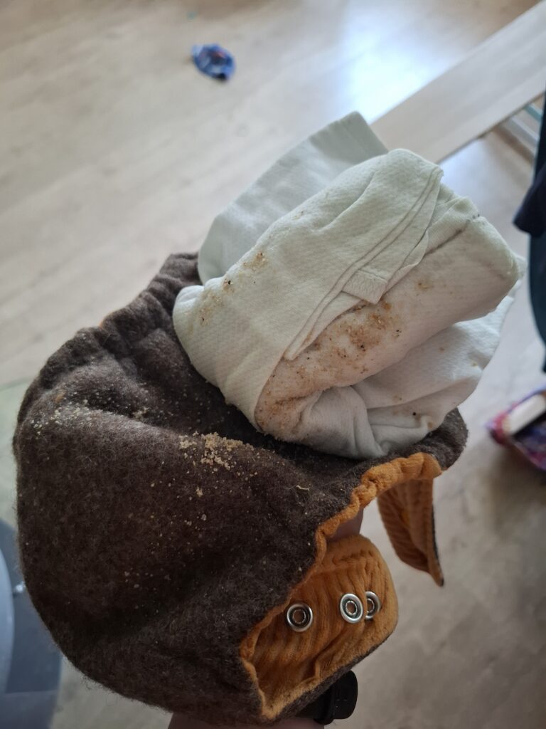 Nahaufnahme einer gebrauchten Wollwindel mit sandiger Einlage in einer Hand, im Hintergrund ein Raum mit Holzfußboden – Beispiel für Stoffwindeln im Alltag.