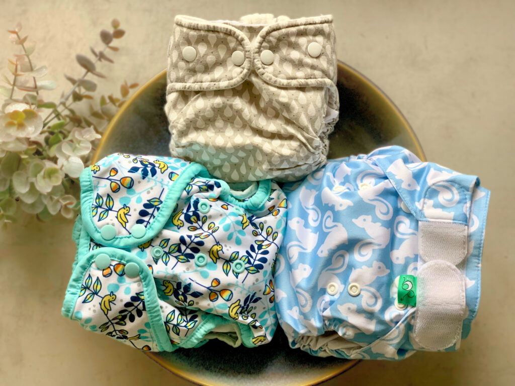 Drei bunte Stoffwindeln liegen in einer Schale: eine graue mit Tropfenmuster, eine weiße mit türkisfarbenem Rand und Zitronenmuster sowie eine hellblaue mit weißen Tiermotiven.